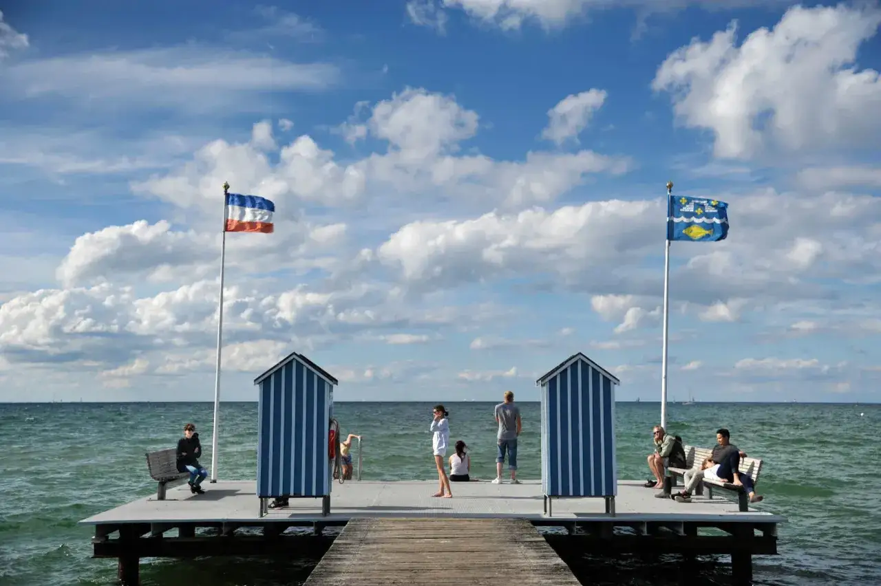 Menschen auf einem Steg an der Ostsee, mit kleinen blauen Strandhäuschen und Fahnen im Wind. Ein schöner Tag am Steinstrand.