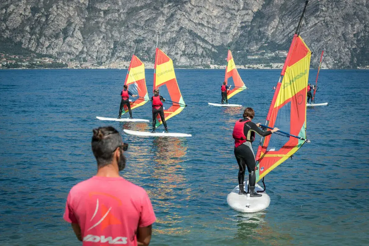 Windsurfer üben am Gardasee. Mehrere Personen auf Surfbrettern mit bunten Segeln gleiten über das blaue Wasser vor einer Bergkulisse.