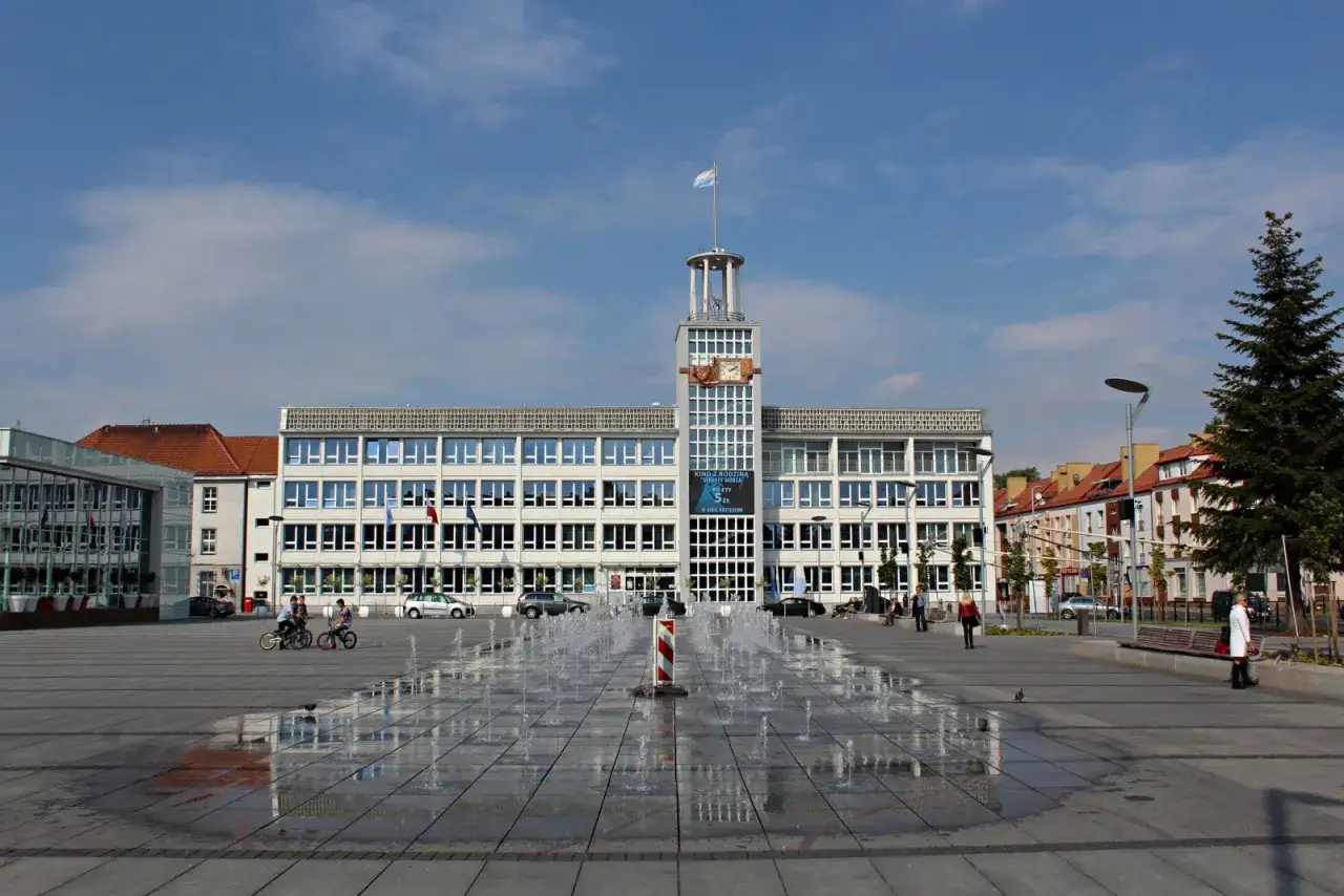 Plac miejski w Koszalinie, z fontannami i ratuszem w tle. Widać ludzi spacerujących i dzieci na rowerach.