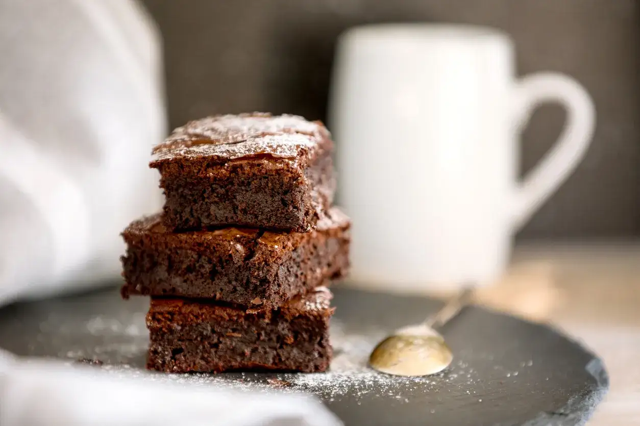 Stos puszystych ciast brownie z płynną czekoladą, posypanych cukrem pudrem, obok łyżeczki i białego kubka.
