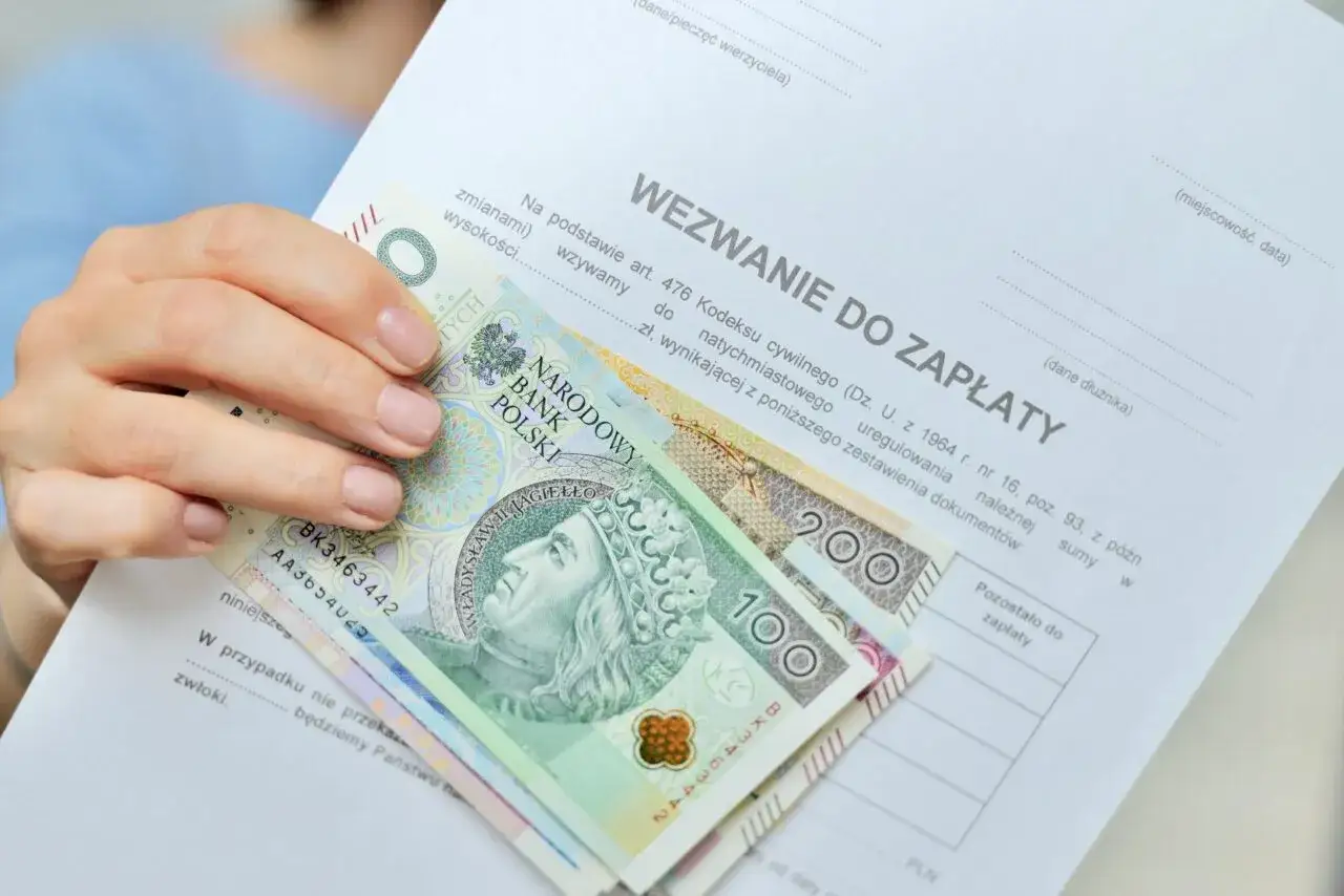 Dłoń trzyma wezwanie do zapłaty i polskie banknoty 100 i 200 zł. Dokument zawiera informację o zawiadomieniu o uchyleniu zajęcia.