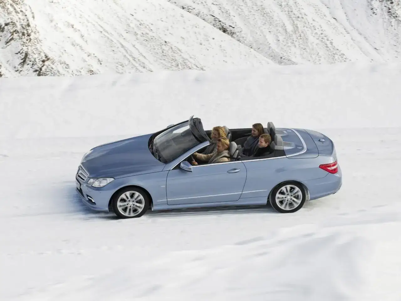 Ein blaues Cabrio fährt durch eine verschneite Landschaft. Vier Personen genießen die Fahrt im Freien, trotz des Winters.