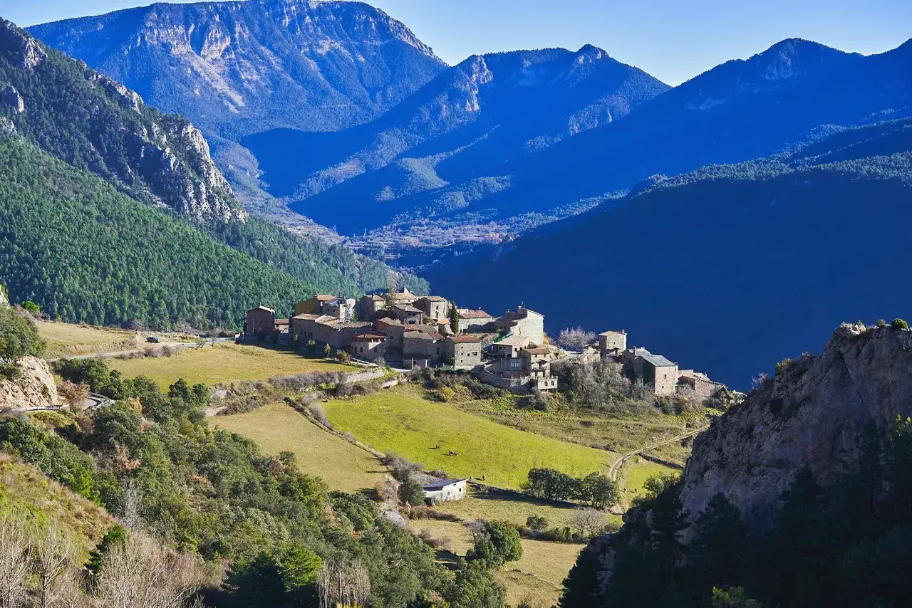 Pueblo de piedra en el **Vall de la Vansa**, rodeado de montañas azules y verdes prados.