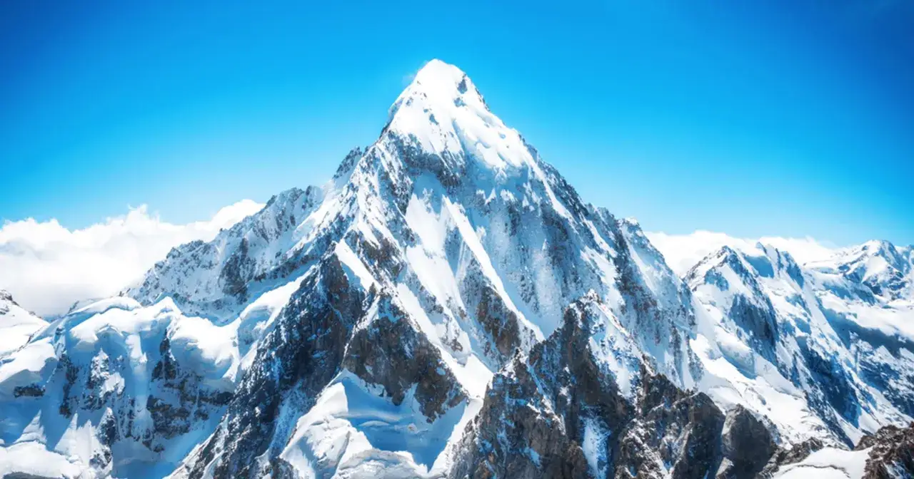 Majestatyczny, ośnieżony szczyt, kt&oacute;ry może być najwyższą g&oacute;rą na świecie, g&oacute;ruje nad innymi, skalistymi wierzchołkami w czystym, błękitnym niebie.
