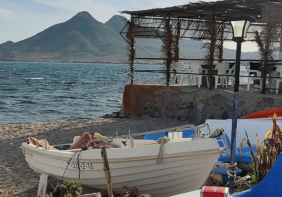 Barca en la playa, con montañas al fondo. Un lugar ideal para saber donde comer en Cabo de Gata, con mesas y sillas bajo un toldo.