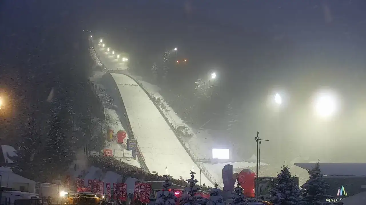 Nocna skocznia narciarska w Zakopanem. Oświetlona trasa czeka na skoczków, a wokół tłumy kibiców.