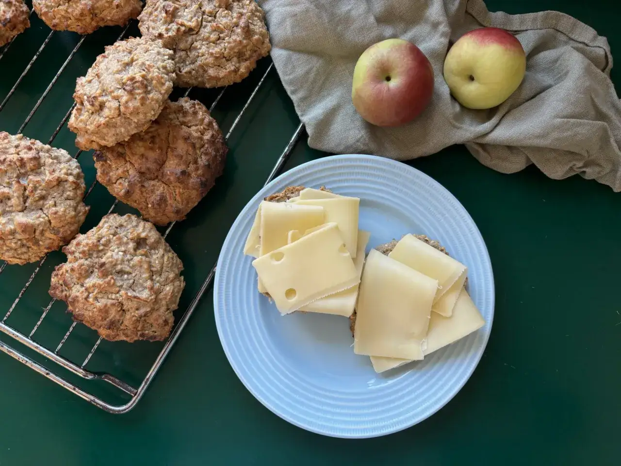 Nybakade äppelscones på ett galler, bredvid två äpplen och en smörgås med ost.
