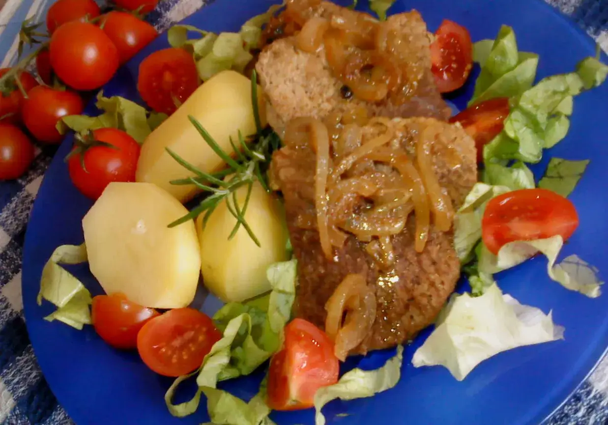Pyszny stek wieprzowy z cebulką, ziemniakami, pomidorkami i sałatą na niebieskim talerzu.