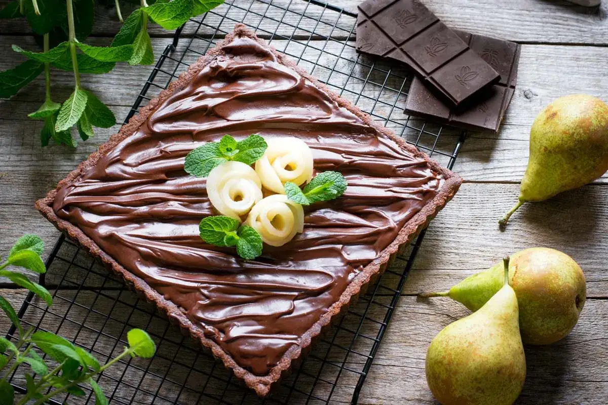 Torta quadrata con crema al cioccolato, decorata con rose di pera e menta. Accanto, pere fresche e tavolette di cioccolato fondente.