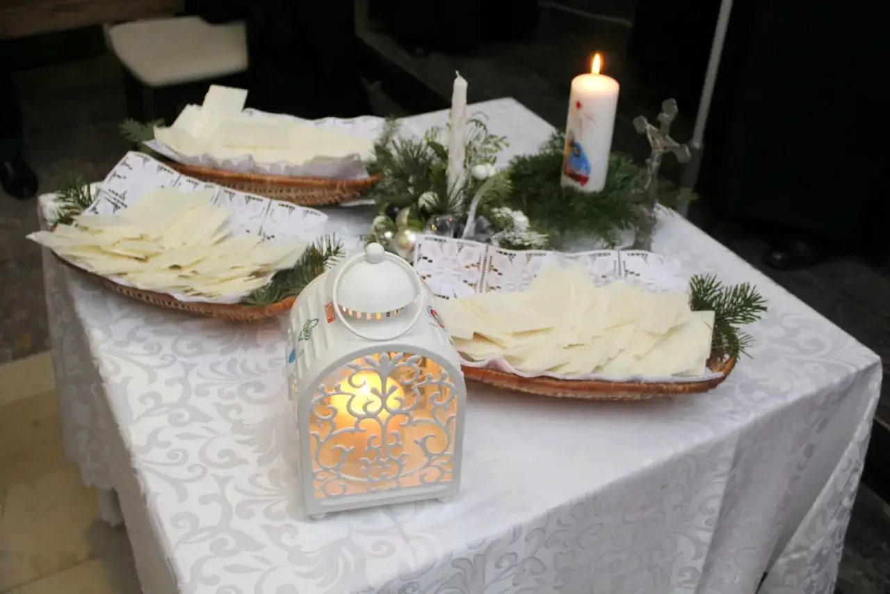 Wigilia. Na stole, przykrytym białym obrusem, stoją koszyczki z opłatkami, ozdobione gałązkami choinki. Płonie świeca, a obok niej lśni metalowy krzyżyk.