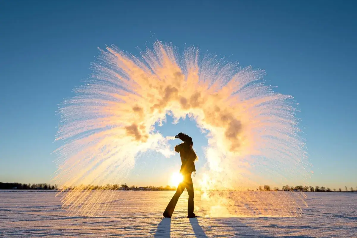 Person kastar varmt vatten i luften i kyla. Vattnet fryser till ispartiklar som lyser i solnedg&aring;ngen. En perfekt slutartid kamera f&aring;ngar &ouml;gonblicket.