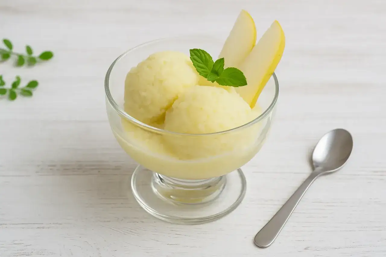 Ein Glas mit cremigem birnen eis, garniert mit Birnenspalten und Minze. Daneben liegt ein Löffel.