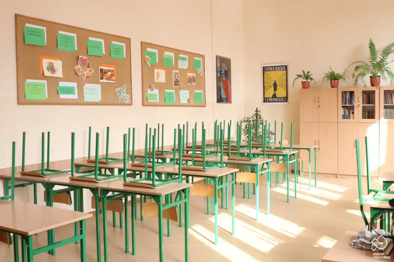 Puste ławki i krzesła odwrócone do góry nogami w sali lekcyjnej. Na ścianach wiszą plakaty i prace uczniów, co to jest szkoła.