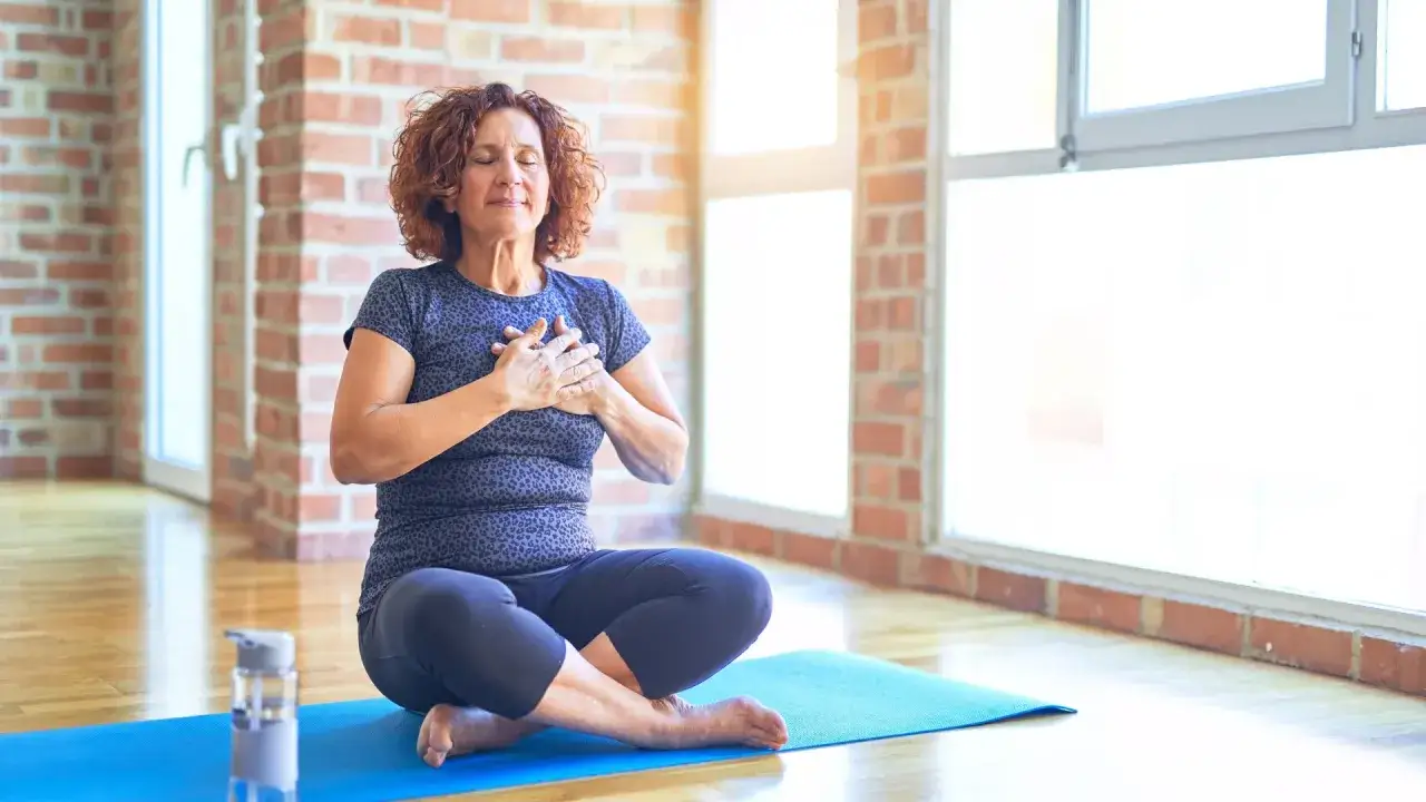 Kvinna med slutna ögon sitter i lotusställning på en yogamatta, med händerna över hjärtat. Hon utövar yoga mot ångest och finner lugn.