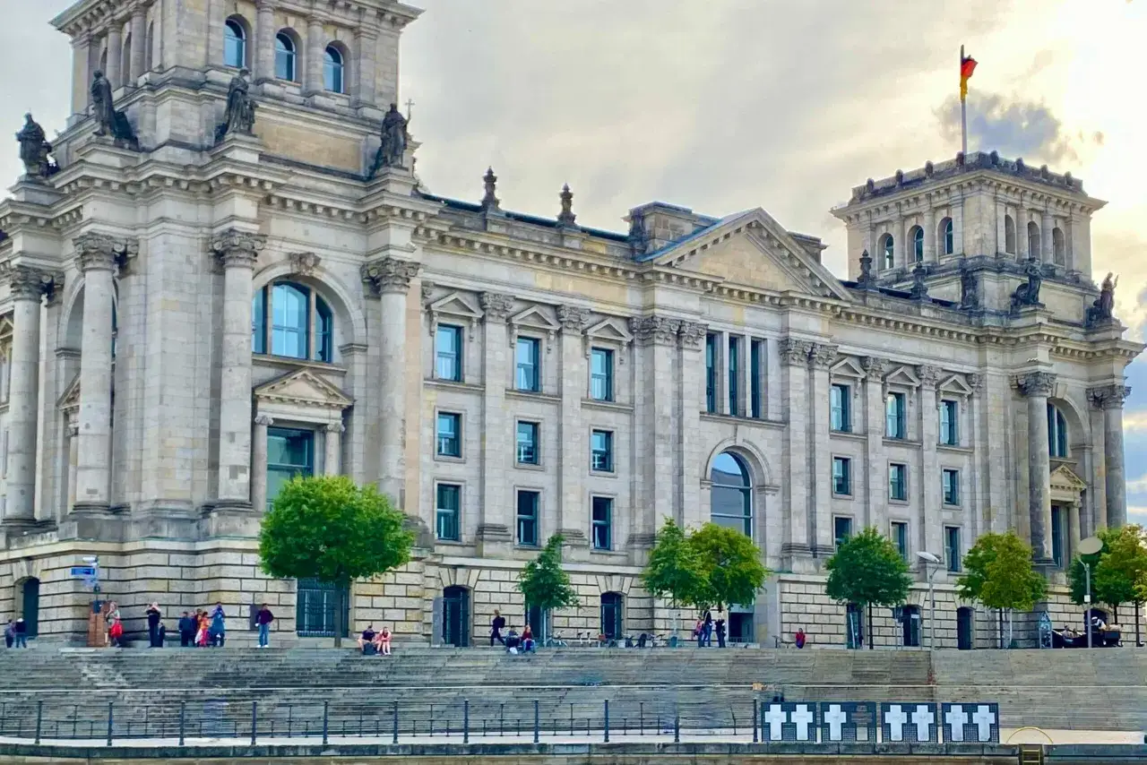 Das Reichstagsgeb&auml;ude im Regierungsviertel Berlin, mit deutscher Flagge und Besuchern am Wasser.