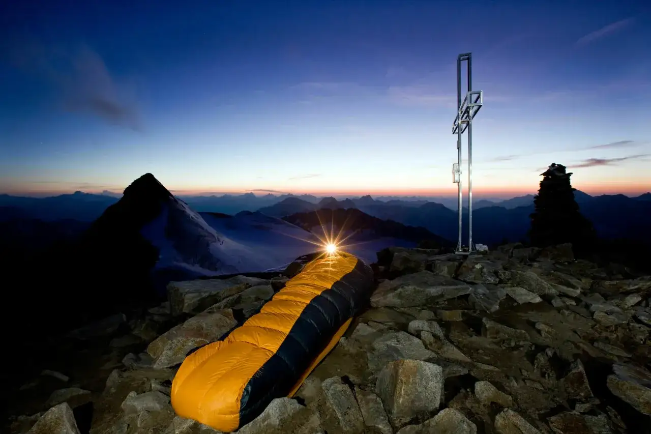 Ein Biwak auf einem Berggipfel bei Sonnenaufgang. Ein leuchtender Schlafsack liegt auf Felsen neben einem Gipfelkreuz. Die wahre Bedeutung von Biwak: Abenteuer und Freiheit.