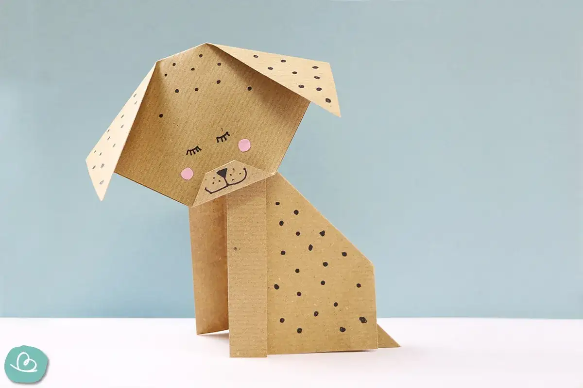 Ein süßer **falten hund** aus braunem Papier mit schwarzen Punkten und rosa Wangen sitzt auf einem weißen Tisch vor hellblauem Hintergrund.