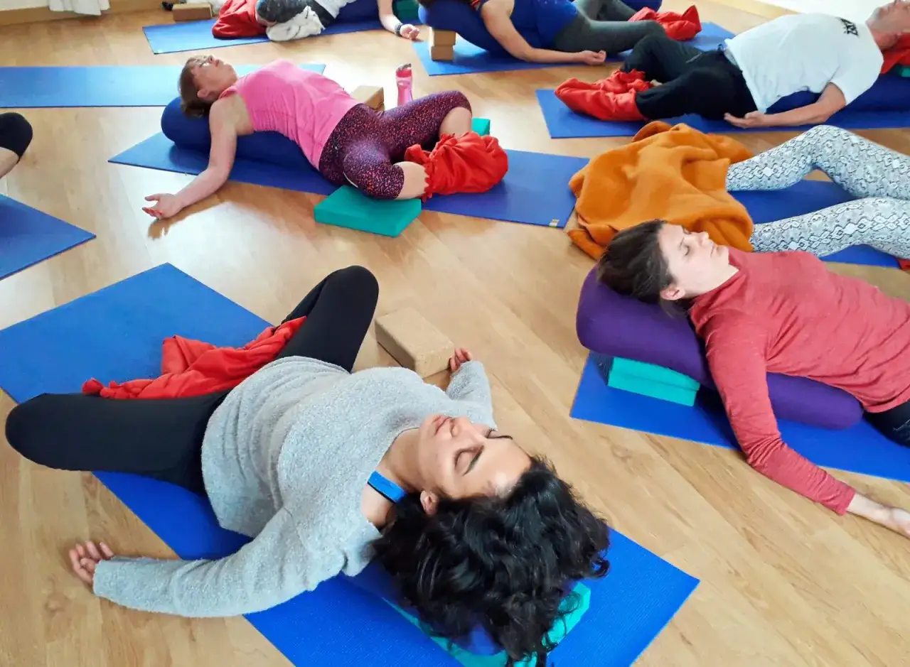 Människor vilar på yogamattor, en del med stöd av bolster och klossar, i en lugn stund av somatisk yoga.