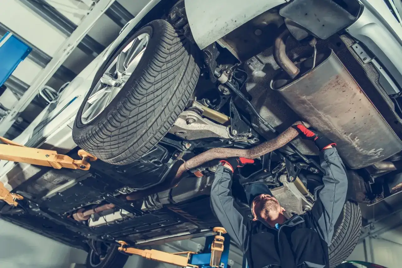 Mechanik w rękawiczkach naprawia wydech samochodu na podnośniku. Dba o to, by luz na kierownicy nie przeszkadzał w bezpiecznej jeździe.