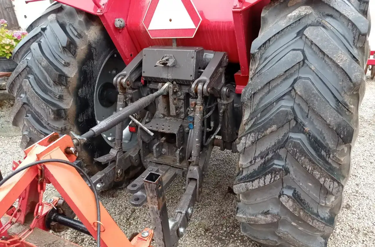 Czerwony traktor z dużymi, brudnymi oponami. Widać jego tylny podnośnik i zaczep. Może zaraz ruszy, ale obawiam się buksowania kół na żwirze.
