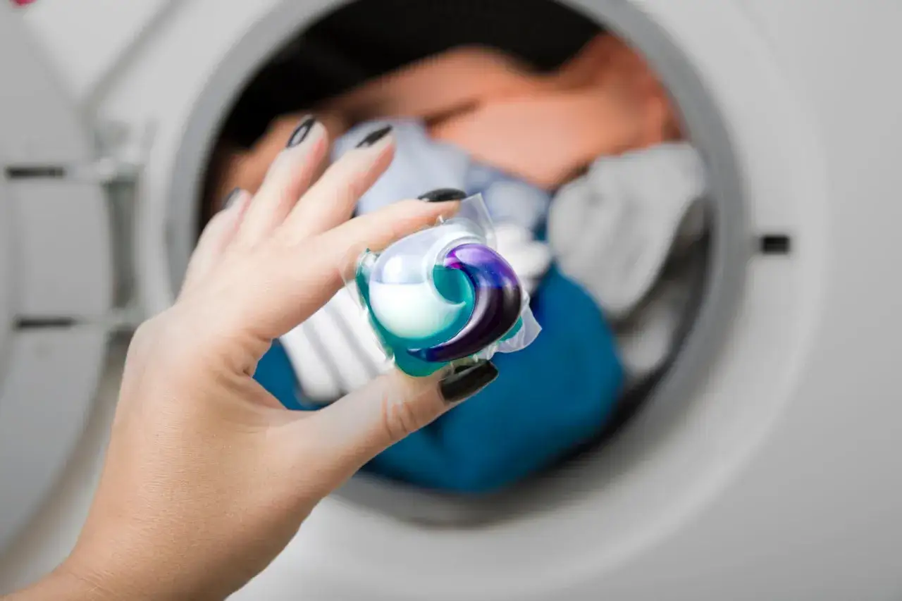 Mano con unghie nere tiene una capsula detersivo colorata, pronta per essere inserita nella lavatrice. Attenzione, le capsule rovinano la lavatrice se usate male.