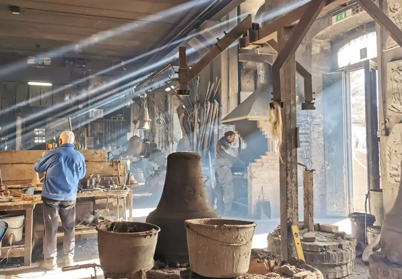 Artigiani al lavoro in una fonderia di **Agnone campane**. Raggi di sole illuminano il fumo e gli attrezzi, mentre le campane attendono di prendere forma.