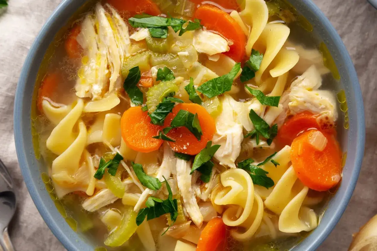 Rozgrzewająca zupa na rosole z makaronem, kawałkami kurczaka, marchewką i selerem, posypana natką pietruszki.