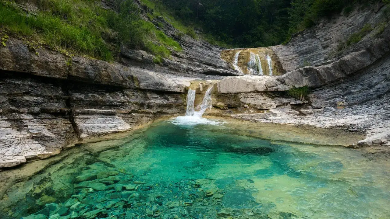 Cascada en un **valle de hecho** con agua turquesa cristalina y rocas estratificadas.