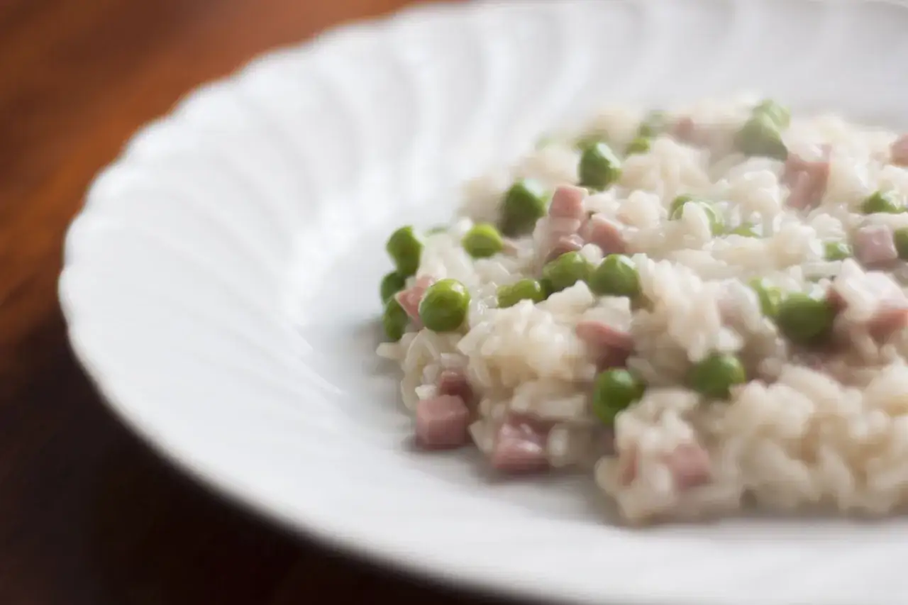 Un cremoso riso con piselli e prosciutto, servito su un piatto bianco con un bordo ondulato.