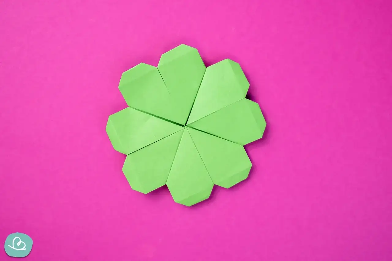 Ein gr&uuml;ner Origami-Gl&uuml;cksbringer, der wie ein Kleeblatt aussieht, liegt auf einem pinken Hintergrund.