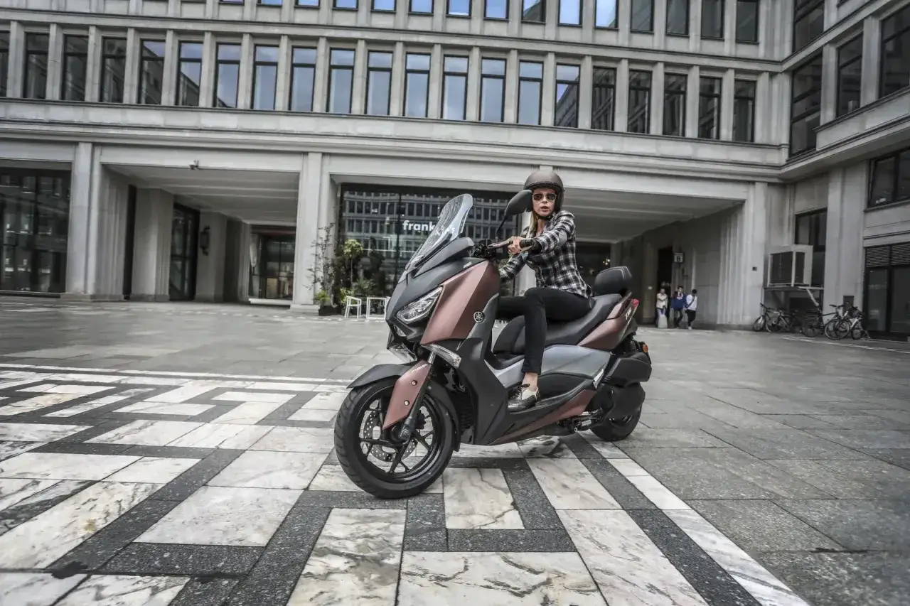 Kobieta w kasku i kratkę na skuterze Yamaha XMAX 300, pozuje na tle nowoczesnego budynku.