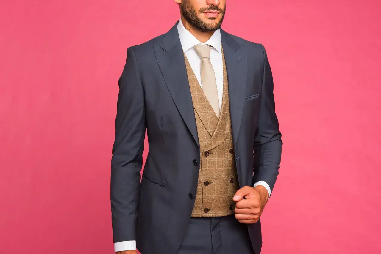 Uomo con abito blu, camicia bianca, cravatta beige e panciotto a quadri marrone. Il panciotto significato è eleganza.