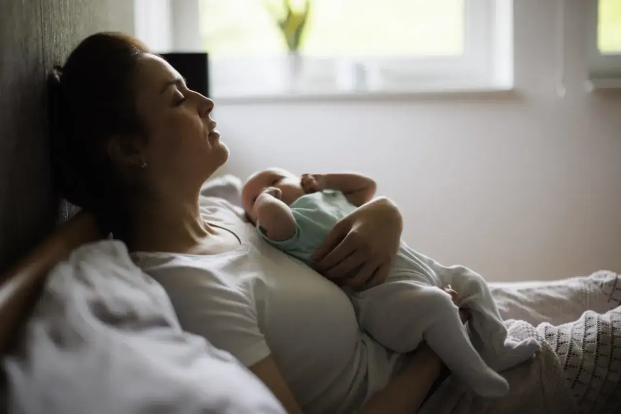 En mamma vilar mot en vägg med sin sovande bebis i famnen. Trots trötthet och en känsla av baby blues, finns det en stilla kärlek i hennes ansikte.