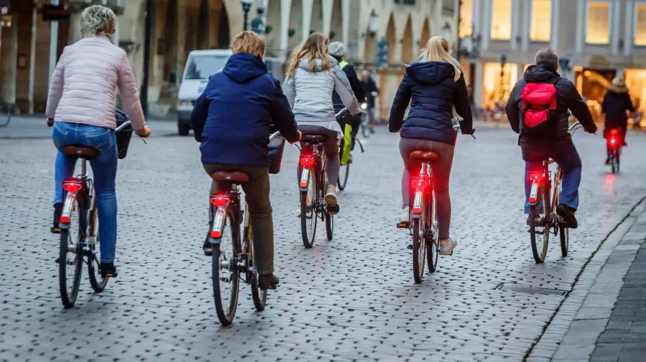 Mehrere Personen fahren nebeneinander Fahrrad auf einer Kopfsteinpflasterstra&szlig;e. Die R&uuml;cklichter ihrer Fahrr&auml;der leuchten rot.