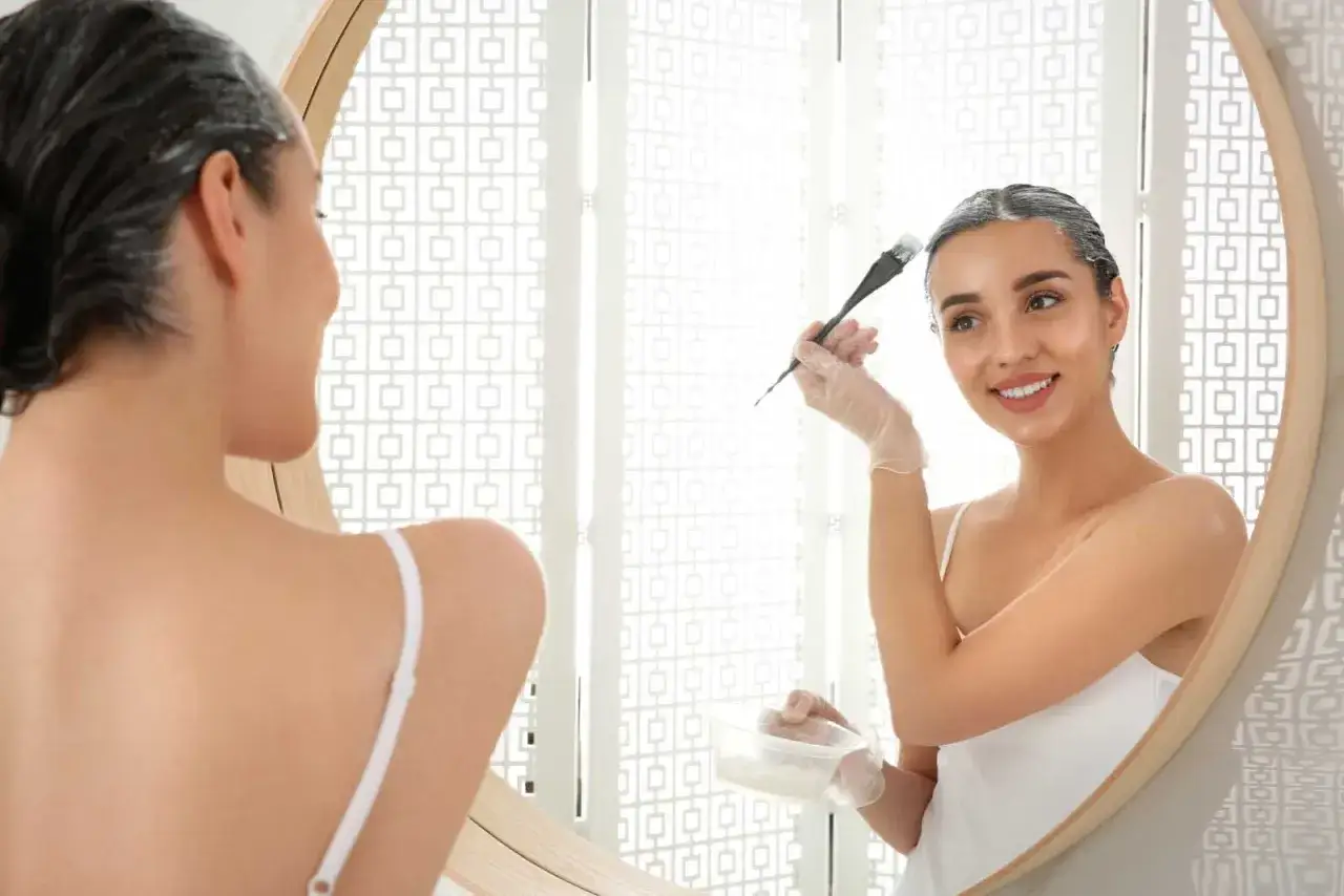 Mujer sonriente se aplica tinta en el cabello en casa, reflejada en un espejo.