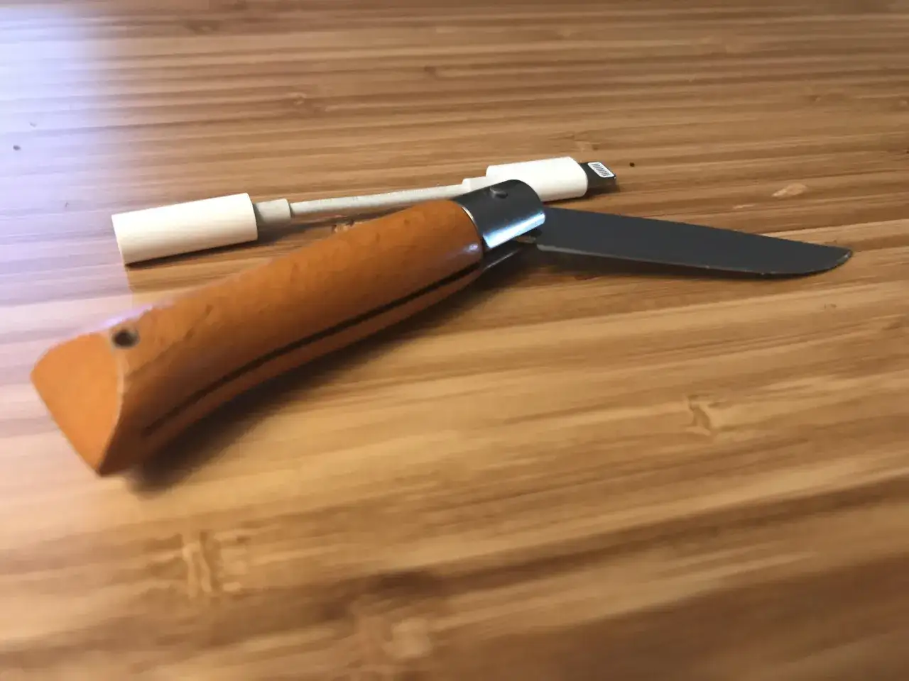 Navaja de bolsillo con mango de madera y hoja de metal, junto a un adaptador de cable blanco. Una daga t&aacute;ctica lista para cualquier situaci&oacute;n.