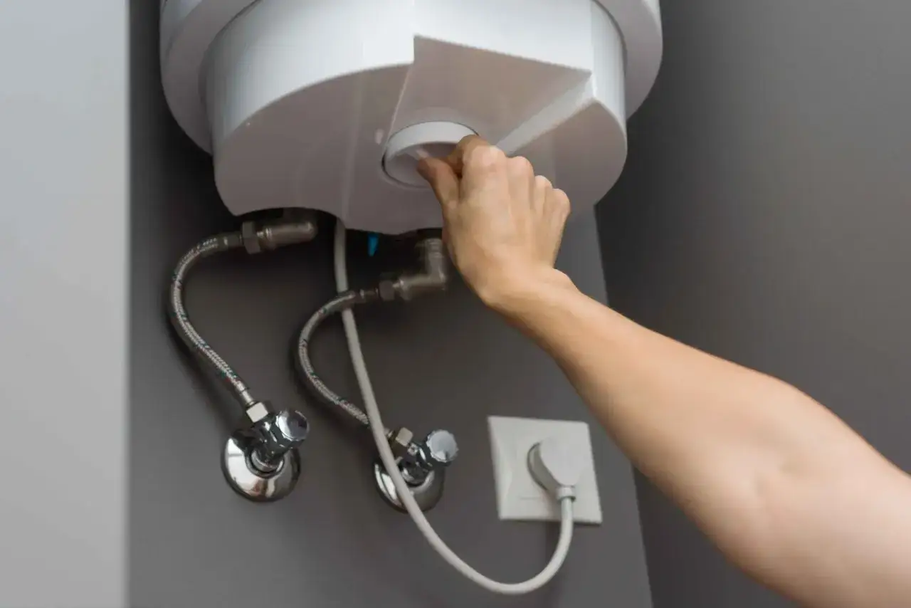 Mano ajustando el dial de un termo el&eacute;ctrico reci&eacute;n instalado. Conexiones de agua y enchufe visibles.