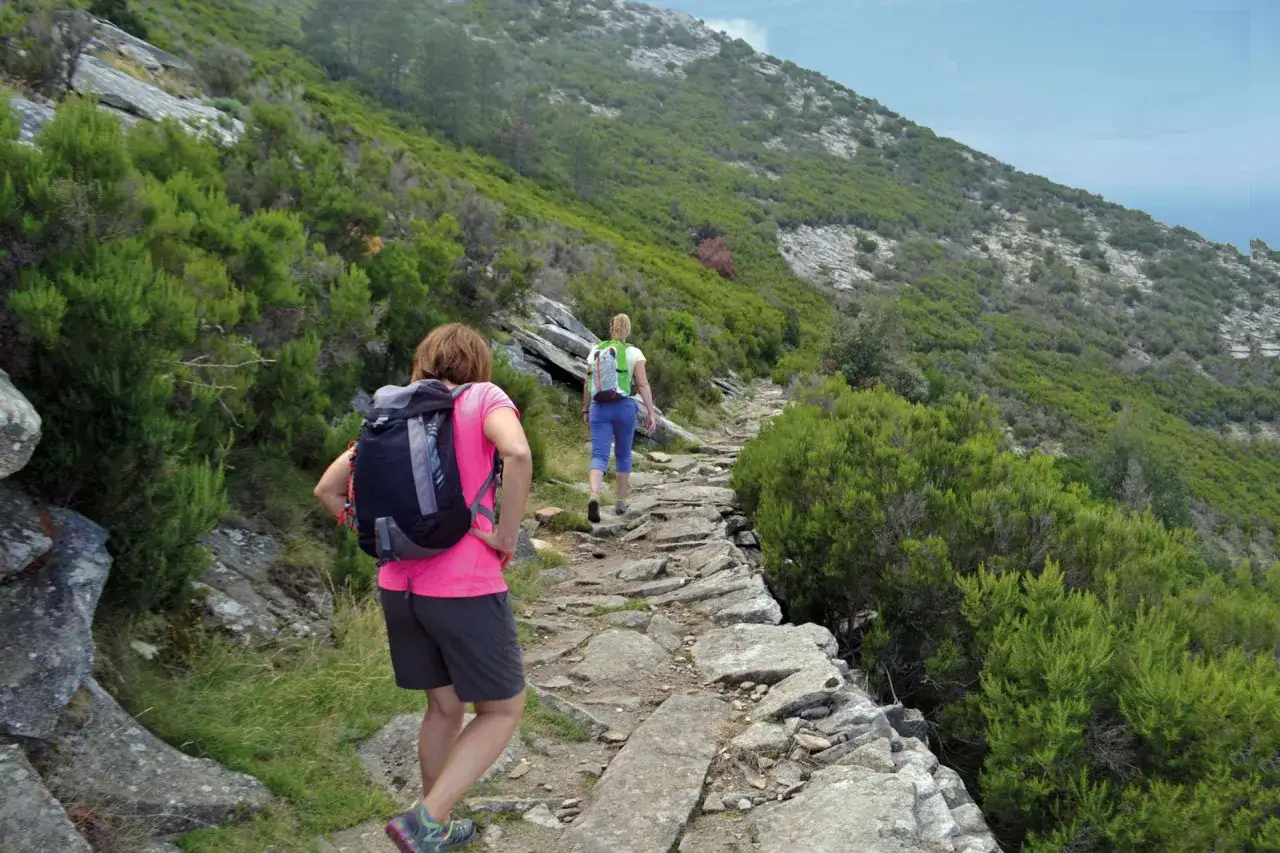 Due escursioniste percorrono un sentiero roccioso su un fianco della montagna, immersi nella macchia mediterranea. La via dell'essenza sull'Isola d'Elba offre panorami mozzafiato.
