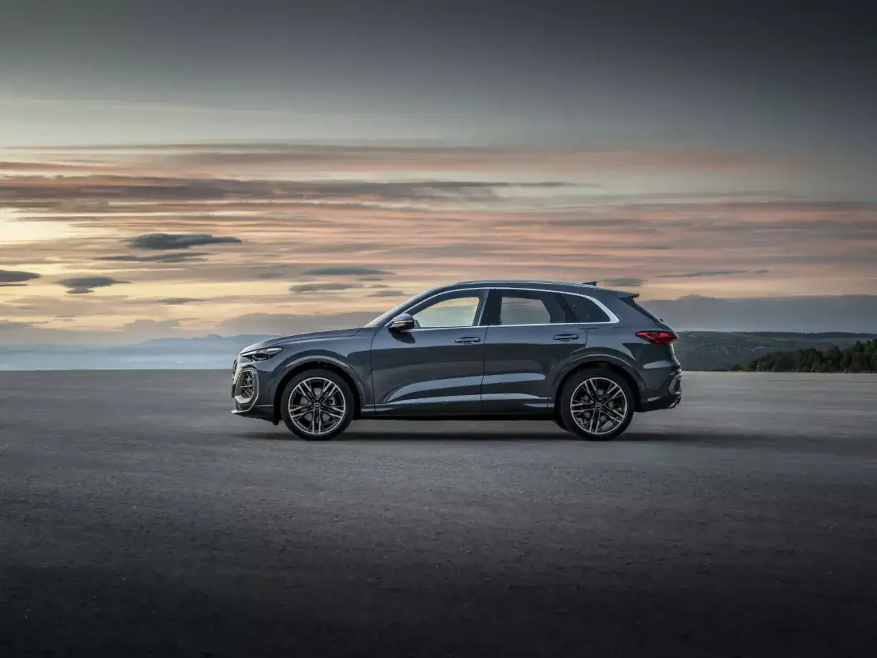 Szary Audi Q5, najnowsza generacja, prezentuje się na tle zachodzącego słońca. Elegancka sylwetka SUV-a na pustej drodze.