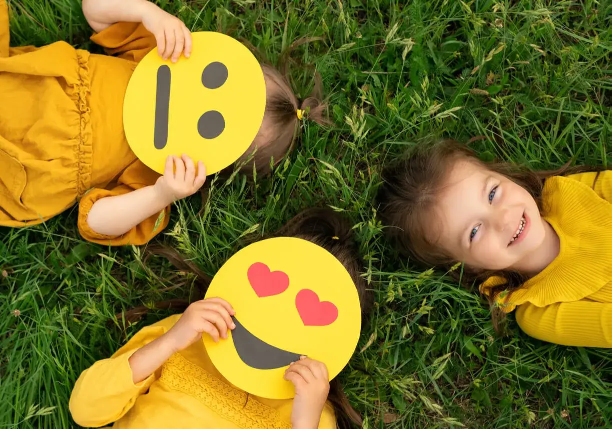 Niñas jugando con caras de emojis para imprimir, expresando alegría y amor en la hierba.