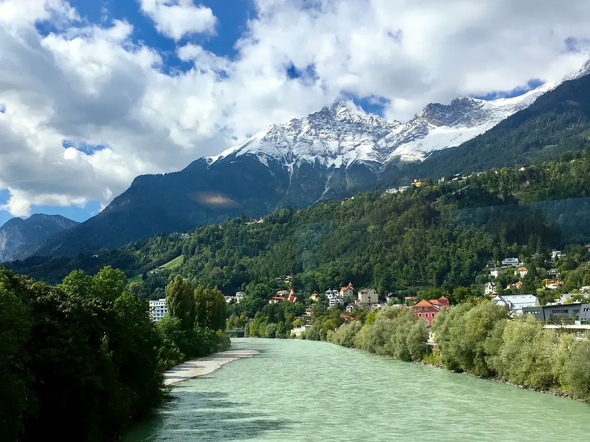 Grön flod flyter förbi ett grönt berg med snöklädda toppar. Hus klättrar uppför sluttningen, en idyllisk vy som påminner om Innsbruck skidåkning.