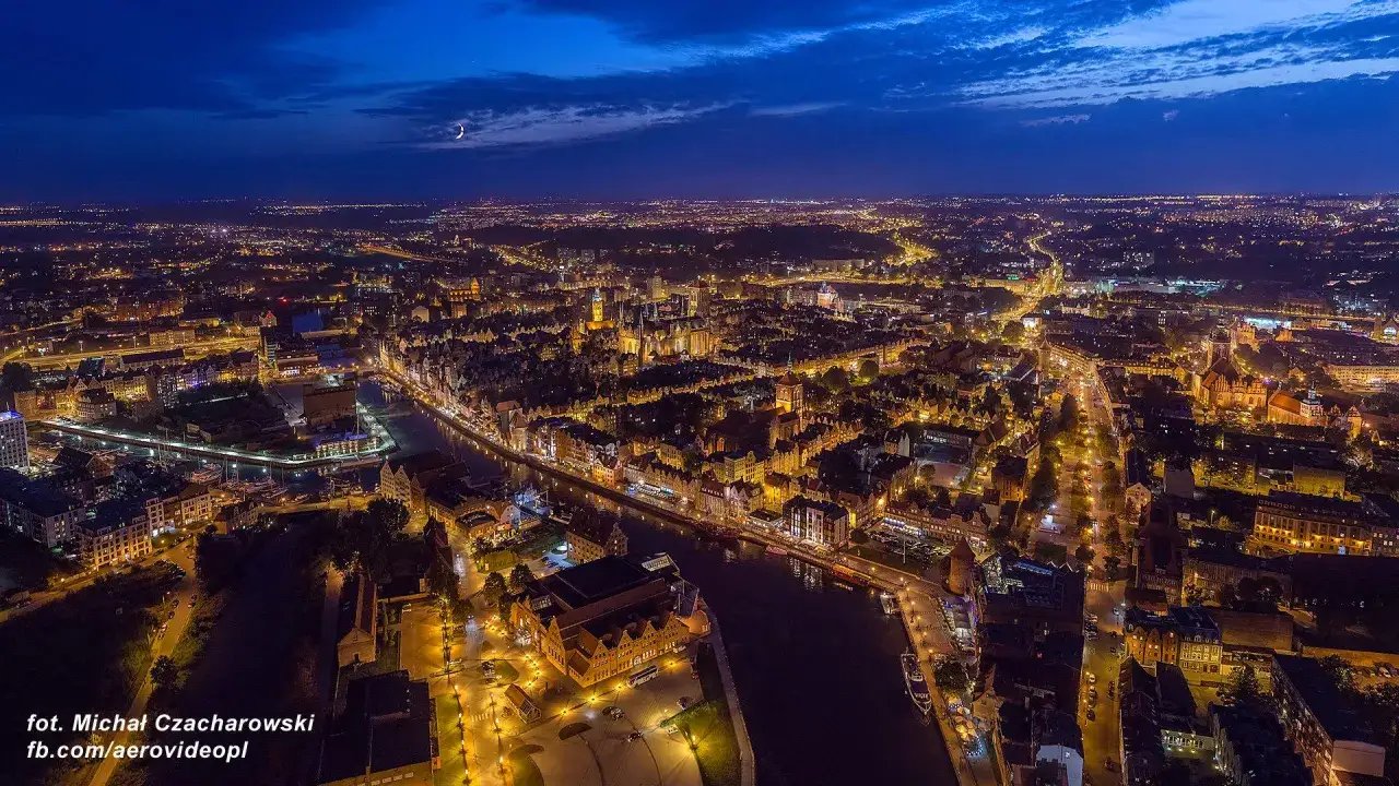 Nocny widok na rozświetlone miasto, rzekę i łodzie. W oddali widać księżyc. To piękny fragment m trójmiasto.