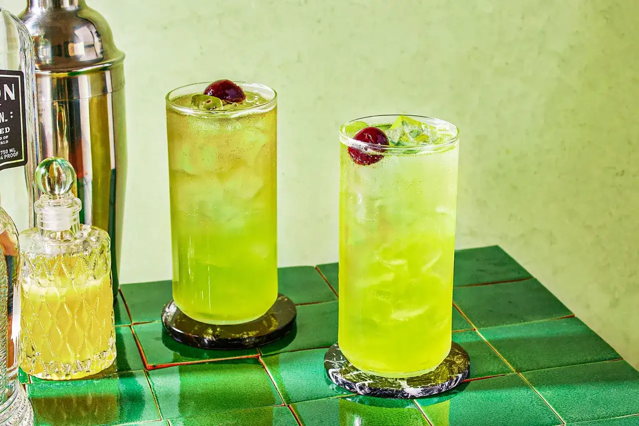 Dwa orzeźwiające drinki Tokyo Iced Tea z wiśniami i lodem, obok shakera i butelki z alkoholem.