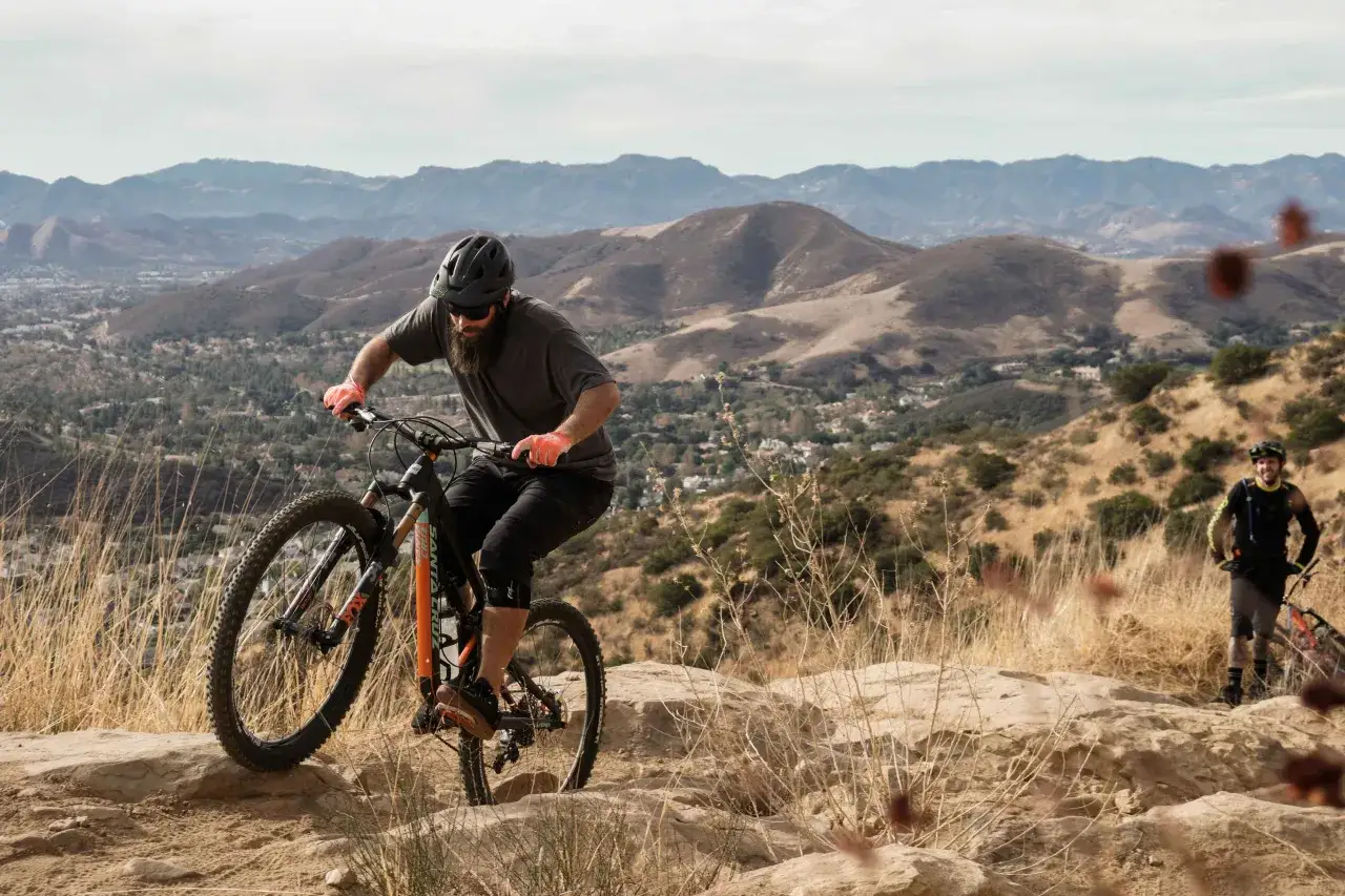 A man with a beard and helmet enjoys mountain biking in Los Angeles, with a friend in the background.