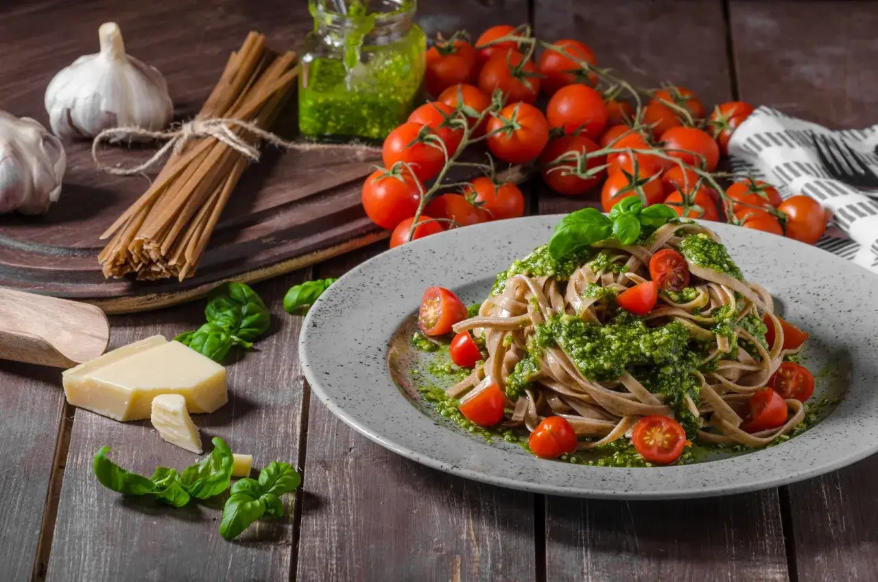 Pyszny włoski obiad: makaron z pesto, pomidorkami koktajlowymi i bazylią. Obok czosnek, ser i świeże składniki.