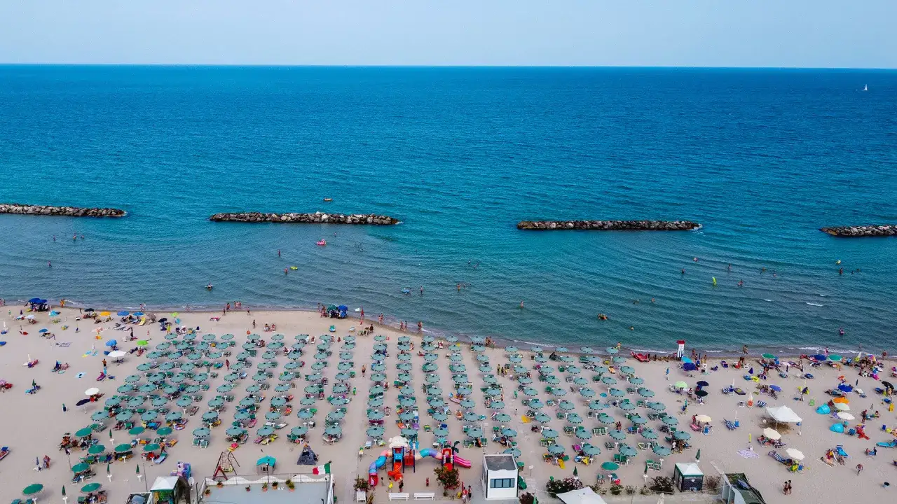 Spiaggia affollata con ombrelloni verdi e blu, bagnanti in mare e frangiflutti in pietra. Un'immagine di un perfetto giorno di **fermo mare**.