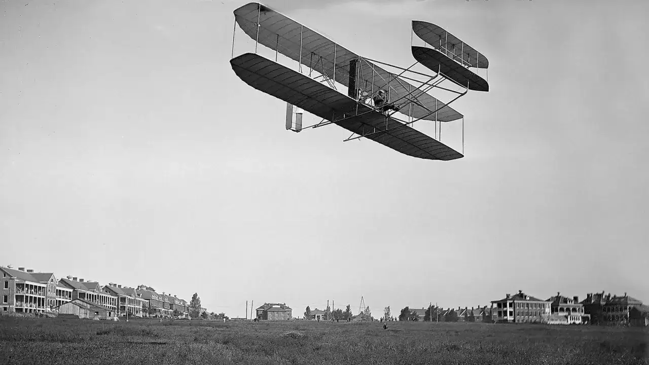 Das Flugzeug der Gebrüder Wright fliegt über ein Feld mit Gebäuden im Hintergrund.