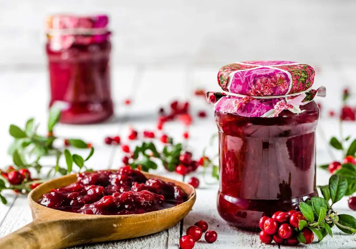 Słoik z domową żurawiną, obok drewniana łyżka pełna konfitury i gałązki świeżych owoców.