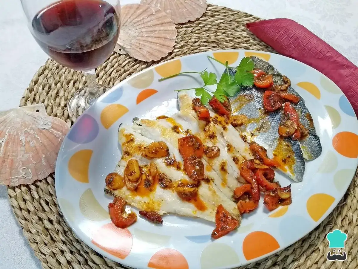 Lubina a la bilbaina servida en un plato con pimientos y ajo, acompañada de una copa de vino tinto y conchas marinas.