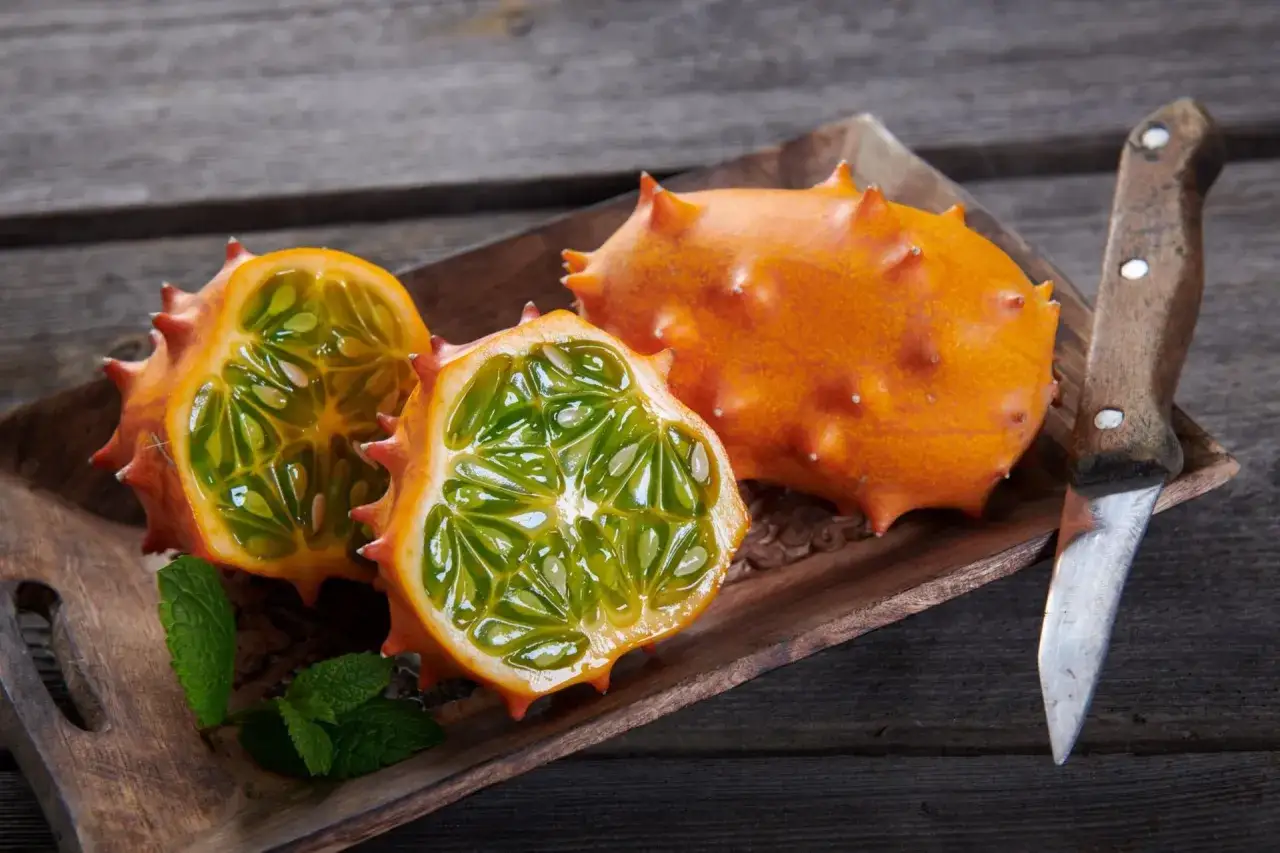 Kiwano jak jeść? Na drewnianej desce leżą dwa przekrojone, pomarańczowe owoce kiwano z zielonym, galaretowatym miąższem i pestkami. Obok leży nóż.